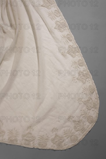 Dress with train, Ludwika Queen of Prussia; 1776–1810 — souvenirs. Dress of white muslin, high waist, with a train, embroidered. Deep-cut square neckline, cinched at the front with a drawstring. Three-quarter sleeves, gathered at the top, flared at the bottom, set-in. Skirt sewn to the bodice, narrow at the front, fuller at the back, with a long train. Front decorated with embroidery: between two rows of stylized flower baskets are small scatterings of polka dots. Sleeves feature embroidered inserts with smaller geometric patterns. Hem and train decorated with stylized flower baskets. Trains clothing, geometric ornaments, Empire style, basket with flowers ornament. Early 19th century, Silesia. Women's clothing. Length 131 cm front, length 230 cm back, circumference 408 cm hem., A pale off-white fabric with soft folds and a scalloped curved edge sits against a muted gray background; along the fabric's border there is a repeating raised floral and heart-like embroidered pattern in a matc...