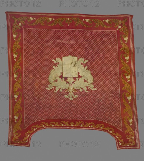 Saddle pad — kilim in a near-square form with slightly elongated upper corners. Main field: rhomboidal mesh ornament with small meshes; central rectangular shield depicting the Boncza unicorn coat of arms, framed at the top by wide and long labres. Surrounded by a border with acanthus leaf motifs arranged in a tendril. Colors: dark red background; ornament in cream, celadon, and tobacco. Date: 1830s. Place of origin: Poland. Material: fabrics. Dimensions: height 248 cm, width 235 cm., A rectangular textile with a dominant red field and a patterned lattice of lighter lines filling the central area, bordered by a wide red band decorated with scrolling floral and acanthus motifs in shades of gold, ochre, and pale cream, with the border motifs repeated along all sides; in the center sits an ornate symmetrical emblem formed of pale cream and gold embroidered foliage and scrollwork surrounding a plain pale rectangular panel, and the lower edge of the textile has a curved cutout with matching...
