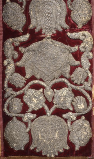 Fragment of embroidered fabric, embroidery, coats of arms, Poland culture, flowers, Jastrzebiec coat of arms, plant ornament. A dark cherry velvet belt embroidered with motifs of large flower heads entwined with branches with smaller flowers and leaves. Among the flowers are stylized tulips, roses, bellflowers, and carnations. At the bottom, under the crown, the Jastrzebiec coat of arms. 17th/18th century, Poland, fabrics, length 237 cm, width 31 cm., A vertical textile panel showing an ornate embroidered pattern on a deep red velvet background, with a repeating symmetrical design of large floral and leaf-like shapes worked in metallic silver thread; the silver embroidery forms a central stacked motif resembling layered petals or scales, surrounded by curling vine-like elements and several stylized blossoms and buds, all rendered with dense, textured stitching; the velvet shows slight wear and variations in red tone, and the silver thread has areas of tarnish and fraying that reveal li...