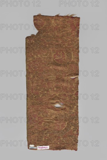 Fragment of a brocade with a large repeat, probably a net composition, with stylized plant motifs pomegranate. Brocaded with gold thread in an S braid on a yellow silk core fringed, accented with drawn gold thread. Colors: cherry red background, yellow and gold ornament. Cherry warp; yellow, gold, and natural-colored silk wefts raw silk weft. Green selvedge. Pattern drawing difficult to read due to worn brocades and background parts. Selection width: 0.4–0.5 cm. 16th century, Spain, fabrics. Height 71.5 cm, width 27.5 cm. Fragment of silk fabric; patterned brocade; fabrics with preserved selvedge; fabrics with drawn thread., A long, narrow textile fragment with a worn, uneven edge and a few small holes, showing a woven pattern of swirling motifs; the fabric appears primarily reddish-brown with areas of golden metallic thread and some beige and pale tan from the worn fibers, set against a neutral gray background.