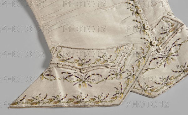 Men's waistcoat of cream silk, embroidered. Straight cut, without a collar; trapezoidally cut hems; low-set pockets covered with flaps. Fastened to the neck with 12 covered and embroidered buttons; identical buttons under the pocket flaps. Embroidery decorates the edges at the closure, the pocket flaps, and the flaps under the pockets. Small floral garlands and tied bundles of grass. Embroidery with satin stitch. Predominant colors: yellow, green, and dark purple. Laced back, silk lining. Fabric-covered and embroidered buttons. 1780s, Silesia Europe. Length 70 cm., plant motifs; garlands; clothing; men's clothing; waistcoat; garment; repossession provenance., The image shows a section of a pale cream-colored garment with a smooth, slightly shiny fabric and decorative embroidery. There is a pointed flap and a folded edge with small rounded covered buttons along a seam. The embroidery forms repeating floral and leafy motifs in muted yellow, olive green, deep purple, and a pale off-white,...