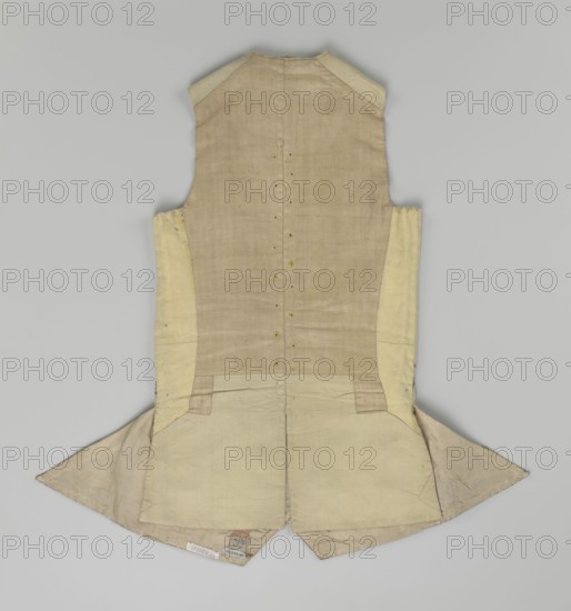 Vest, Floral and plant ornaments on brocaded silk fabric. Predominant ornamental colors: silver, plum, green, and raspberry. Front with motifs arranged around the edges; fastened with 12 buttons embroidered with gold threads; no collar. Side flaps with flapped pockets, three buttons under the flaps as for the closure. Back: bottom and sides of fabric as on the front, cut-out pieces plain without brocade. Cream silk lining. 1740–1760, England fabric possibly from Silesia. Garment; length 74 cm., A sleeveless tailored garment laid flat, showing the back with a fitted waist and two flared skirt-like panels at the bottom; the fabric appears in shades of beige, cream, and light tan with visible stitching lines, seam edges, and small reinforcement patches; there are a few small puncture-like markings along the center back seam and several subtle creases and worn areas on the skirt panels; the shoulders have slightly darker beige patches and the interior lining visible at the bottom edge is a...