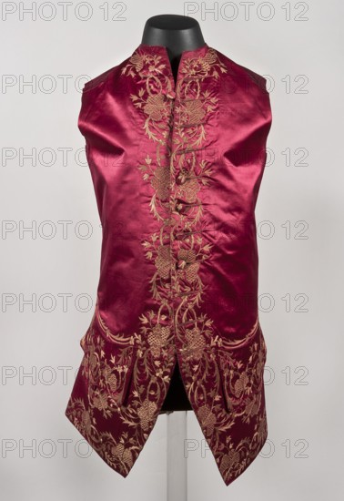 Vest, Men's vest made of cherry satin, embroidered. Straight cut, sleeveless and collarless. Diagonal hemline, side pockets covered with large flaps, three covered buttons under each flap. Fastened to the neck with 11 covered and embroidered buttons. 1740–1770, Silesia. Length 78 cm., floral ornaments., A sleeveless garment displayed on a black mannequin form, made of shiny deep red fabric with intricate gold floral embroidery running down the center front and along the lower edges; the embroidery forms looping vines and clustered flower motifs, and the garment has a high collar and a front opening that splits at the hem into two pointed panels, showing texture variations and gentle folds in the fabric against a pale neutral background.