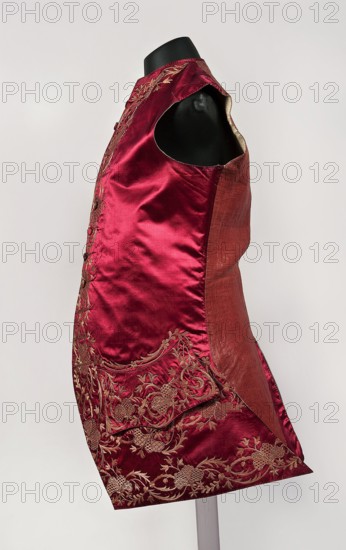Men's vest made of cherry satin, embroidered. Straight cut, sleeveless and collarless. Diagonal hemline, side pockets covered with large flaps, three covered buttons under each flap. Fastened to the neck with 11 covered and embroidered buttons. Floral ornaments. Made in Silesia, 1740–1770. Length 78 cm., provenance includes repossession., A sleeveless garment displayed on a black mannequin torso, made of a shiny deep red fabric with a matte reddish-brown side panel; the red fabric has ornate embroidered floral motifs in a metallic gold thread near the lower and side areas, and there is decorative embroidery along the front edge; the garment shows a smooth satiny texture with some visible folds and seams, and the background is a pale off-white.