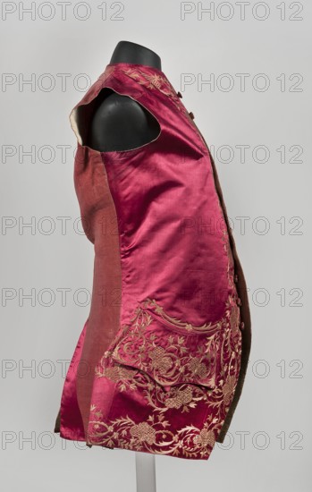 Men's vest made of cherry satin, embroidered. Straight cut, without sleeves or collar. Diagonal hemline, side pockets covered with large flaps, three covered buttons under each flap. Fastened to the neck with 11 covered and embroidered buttons. 1740–1770. Silesia. Length 78 cm., Floral ornaments. Repossession, A sleeveless garment displayed on a black mannequin torso, made of a shiny deep pinkish-red silk with a slightly darker pinkish-red side panel, featuring ornate embroidered floral and scrolling vine motifs in a light tan-gold thread concentrated near the lower front and pocket flap, with matching embroidered detail near the shoulder, a rounded armhole edge showing a pale lining, and a row of fabric-covered buttons along the front edge, set against a neutral light gray background.