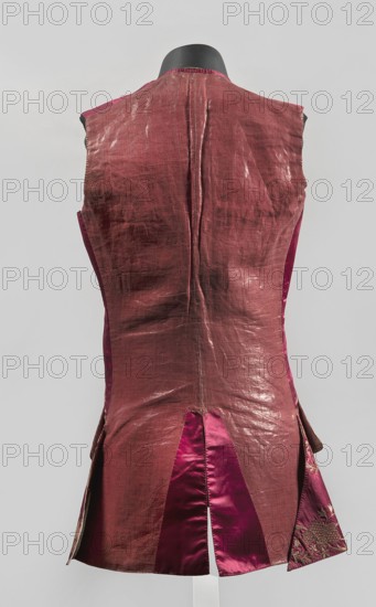 Vest, floral ornaments, men's vest made of cherry satin, embroidered. Straight cut, sleeveless and collarless. Diagonal hemline, side pockets covered with large flaps, three covered buttons under each flap. Fastened to the neck with 11 covered and embroidered buttons. 1740–1770, Silesia, length 78 cm., A sleeveless garment shown from the back on a black display form, made of a muted reddish-brown outer fabric with a slightly shiny, crinkled texture and darker areas of shading; the sides and lower interior panels reveal a richer magenta satin fabric, and one lower side panel shows a patterned magenta fabric with floral embroidery in a darker tone; the background is a pale light gray.