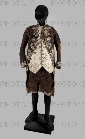 Vest, floral and geometric ornaments. Fastened at the neck with 9 covered and embroidered buttons. Side flaps with pockets underneath. Laced canvas back. 2nd half of the 18th century, Poland. Garment, length 74 cm., A mannequin with a black head and black legs stands on a black rectangular base wearing a brown corduroy long coat and matching knee-length breeches, paired with a cream-colored waistcoat and vest panels that have floral embroidery. The coat features decorative floral appliqués and circular ornamental buttons along the front and cuffs, and the knee hems and inner edges show embroidered trim. The outfit includes black shoes with a low heel and strap details. The visible colors are brown, cream, black, and touches of metallic and pale blue in the embroidered floral decorations, set against a pale gray background.