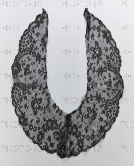 Collar, lace clothing accessory for women's fashion; lace collar with shell-and-lace motifs around the outer serrated edge. Collar is widest at the middle, tapering to elongated ends; inner edges are cut and unsecured. 2nd half of the 19th century. Possible origin: Belgium or France. Materials: lace. Dimensions: width 23 cm, length 81 cm., A delicate lace garment in black laid flat against a light gray background, forming a U-shaped collar with scalloped outer edges and a narrow opening at the top, the lace featuring an intricate floral pattern with clusters of small and larger flower motifs and leafy swirls, the fabric appearing sheer with fine netting between the embroidered designs and subtle variations in texture where the floral embroidery is denser, the edges showing slight irregularities and soft frayed threads in places.