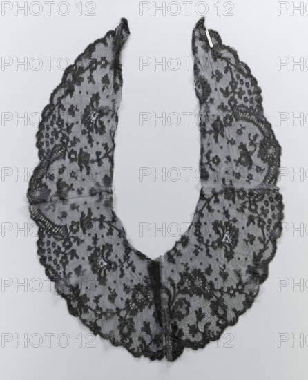 Collar, lace clothing accessory for women's fashion. Around the outer serrated edge are shell-and-lace motifs. The collar is widest in the middle, tapering at the elongated ends. The inner edges are cut and unsecured. 2nd half of the 19th century. Probable origin: Belgium or France. Materials: lace; dimensions: width 23 cm, length 81 cm., A delicate scalloped lace collar in black is laid flat on a pale gray background, forming a U-shape with two elongated curved panels meeting at the bottom; the lace features an ornate floral pattern with clusters of flowers, leaves, and scrolling vines worked into a fine mesh, and the edges are finished with a scalloped trim.