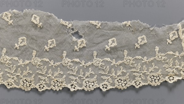 Lace fragment, lace, provenance unknown, plant ornaments, tulle embroidered with twig and floral motifs around the lower edge, and projections of small twigs above. Upper edge cut off, unsecured. 19th century, Europe, lace, width 12 cm, length 56 cm, A length of delicate lace fabric displayed against a gray background, the lace is an off-white or cream color with a fine hexagonal mesh ground; the lower edge features a scalloped border of embroidered floral motifs with open circular rosettes and leafy sprigs, above which runs a garland of small embroidered flowers and loops, and the upper field is scattered with small embroidered diamond-shaped motifs; there are a few irregular worn areas and a small hole near the top center where the mesh is torn, and the overall texture appears lacy and slightly frayed at the torn and outer edges.