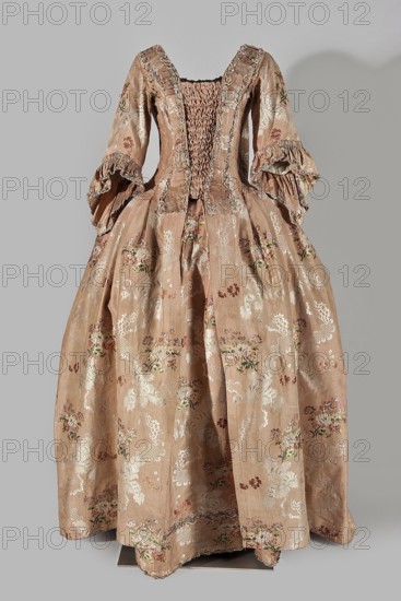 Two-piece robe à la française dress, silk in the colour of dried rose with motifs of colourful flower bouquets and winding white lace ribbons. Overskirt manteau open at the front, with a triple pleat extending almost the entire back and forming a train. Bodice on linen lace with boning, laced at the front; deep square neckline. Narrow elbow-length set-in sleeves trimmed with ruffles of the same fabric. Neckline and bodice edges trimmed with a pleated ruffle decorated with multicolored cord trimmings twisted into tiny flowers. No lining. Skirt gathered with symmetrical pleats, spread to the sides. Patterned fabric; fabric report: height 24.5 cm, width 48.5 cm; width of fabric piece 54 cm. Dated 1760–1765. Origin/attribution: Silesia; possibly France. Length 158 cm. Features: floral ornaments, ribbon ornament, flower bouquet motifs, ruffles; women's 18th-century clothing., A full-length historic-style dress displayed against a plain background, made of a shiny pale pinkish-beige fabric p...