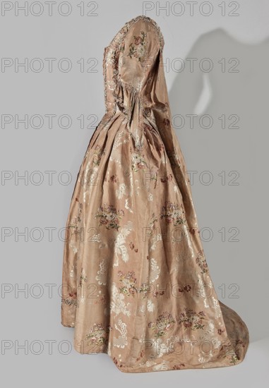 Two-piece robe à la française dress, silk in the colour of dried rose with motifs of colourful flower bouquets and winding white lace ribbons. Overskirt manteau open at the front, with a triple pleat extending almost the entire width of the back, flowing into a train. Bodice on linen lace with boning, laced at the front. Deep, square neckline. Narrow elbow-length set-in sleeves trimmed with ruffles of the same fabric. Neckline and bodice edges trimmed with a pleated ruffle decorated with multicolored cord trimmings twisted into tiny flowers. No lining. Skirt gathered with symmetrical pleats, spread out to the sides. Patterned fabric; floral ornaments, ribbon ornaments, flower bouquets, ruffles. Fabric report: height 24.5 cm, width 48.5 cm; width of fabric piece 54 cm. Dated 1760–1765. Silesia; possible France origin. Length 158 cm., Women's clothing; 18th century., A full-length historical gown displayed on a neutral background, made of a pale peachy-pink silk with a subtle sheen and d...