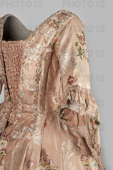 Robe à la française dress, floral ornaments, ribbon ornament, flower bouquets, robe à la française, ruffles clothing decoration, women's clothing, patterned fabrics, 18th century clothing. Two-piece dress made of silk in the color of dried rose with motifs of colorful flower bouquets and winding ribbons of white lace. The overskirt manteau is open at the front, with a triple pleat extending almost the entire width of the back, which flows into a train. The bodice is on linen lace with boning, laced at the front. The neckline is deep, cut in a square shape. Narrow, elbow-length set-in sleeves are trimmed with ruffles made of the same fabric as the dress. The neckline and bodice edges are trimmed with a pleated ruffle, decorated with multicolored cord trimmings twisted into tiny flowers. No lining. The skirt is gathered with symmetrical pleats, spread out to the sides. Fabric report: height 24.5 cm, width 48.5 cm; width of fabric piece: 54 cm. Date: 1760–1765. Place: Silesia; possible or...