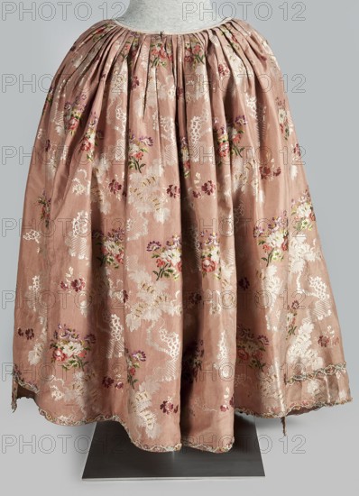 Two-piece robe à la française dress, silk in the colour of dried rose with motifs of colourful flower bouquets and winding white lace ribbons; 1760–1765. Overskirt manteau open at the front, with a triple pleat extending almost the entire width of the back and flowing into a train. Bodice on linen lace with boning, laced at the front; deep square neckline. Narrow elbow-length set-in sleeves trimmed with ruffles of the same fabric. Neckline and bodice edges trimmed with a pleated ruffle decorated with multicolored cord trimmings twisted into tiny flowers. No lining. Skirt gathered with symmetrical pleats, spread to the sides. Patterned fabric; fabric report: height 24.5 cm, width 48.5 cm; width of fabric piece 54 cm; length 158 cm. Floral ornaments, ribbon ornament, flower bouquet motifs, ruffles; women's 18th-century fashion; Silesia; France possible origin., A full-length garment displayed on a mannequin with a gathered waistband, made of a soft pink fabric patterned with floral motif...