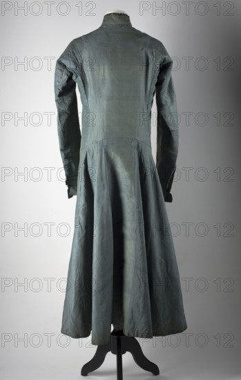 Zupan made of steel-blue grosgrain, long, fastened at the front with leatherwork buttons. Fitted at the top, slightly flared at the bottom. High, stand-up collar. Long, set-in sleeves, finished with turn-up cuffs fastened with hooks. Side pockets. The back is cut with a pole like that of a kontusz, widened from the waist by semi-flared pleats. Lining made of gray linen and indigo-dyed linen, impregnated. Hand-sewn, 18th century, Poland; men's clothing, national costume, length 122 cm, circumference 182 cm bottom. Polish clothing; materials: silk, steel-blue grosgrain, gray and dyed canvas. Creator: unknown Czechman., A long sleeved dress displayed on a mannequin stand, shown from the back, in shades of bluish-green and gray with visible creases and seams; the bodice is fitted with a high collar and long sleeves that taper at the cuffs, and the skirt flares into a flowing, pleated shape reaching toward the floor; the mannequin stand is black and supports the garment against a neutral gr...