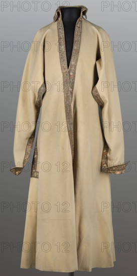 Kontusz from the uniform of the Order of St. Stanislaus. Kontusz of white cloth with lapels of crimson, gold-embroidered grosgrain, edges trimmed with gold braid. Fitted at the top, flared at the bottom, fastened at the front with hooks. The lapels do not meet at the top, creating a triangular neckline, finished with a high, standing, turn-down collar. The back is cut with a central pillar of fabric, widened on the sides from the waist by semi-flared pleats. Pockets in the side seams. Crimson grosgrain brocaded with braided thread and silk threads; gold stripes on a crimson background and miniature sprigs of stylized flowers. Gold galloon Slutsk or Grodno, 1780s, Poland. Clothing, national costume. Length 126 cm; width at bottom 325 cm., A long coat displayed on a black mannequin, made of a pale beige fabric with a slightly textured surface, featuring a standing collar and wide front opening edged with a narrow decorative trim in darker brown and muted green tones with small mottled ar...