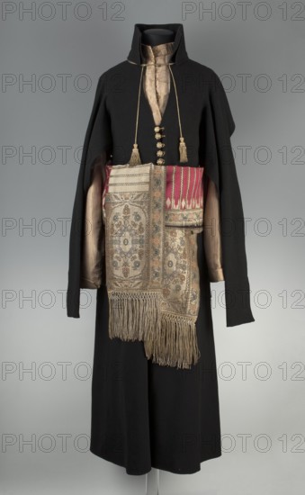 Zupan, Polish men's clothing national costume, long zupan fitted at the top and flared at the bottom. Fastened with hooks and eyes at the neck with a concealed fastening; low stand-up collar. Long set-in tapered sleeves with turn-up cuffs, slit at the bottom and fastened with hooks and eyes. Back cut like a kontusz with a central column of fabric, widened from the waist by semi-flared pleats, adjustable at the top with ribbons. Pockets in the side seams. Silk satin with pearl-brown stripes; back and lining of gray linen canvas. Hand-sewn, 18th/19th century, Poland. Length 118 cm; bottom circumference 243 cm., A mannequin dressed in a long black outer garment with a high collar and long sleeves, layered over a beige striped inner garment; a decorative sash with a patterned panel and long beige fringe hangs around the waist, showing a section with deep pink and cream stripes alongside intricate floral and geometric motifs in muted beige, blue, and brown tones; a pair of long gold cords w...