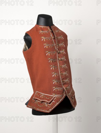 Men's waistcoat, repossession provenance; 2nd half of the 18th century, Silesia. Pink sleeveless, collarless fitted waistcoat, length 70 cm. Fastens at the neck with ten buttons. Bottom sides have pockets with flaps, three buttons under each flap. Skirts cut diagonally at the bottom. Back to the waist in coarse woolen fabric, from the waist down in cloth. Embroidered decoration like outerwear; fabric-covered and embroidered buttons. Embroidery in white, blue, and green untwisted silk yarn. Fustian lining. Hand-sewn. Ornament: flower bouquets, flowers, garlands; floral ornaments; men's clothing/fashion; waistcoat., A sleeveless reddish-brown vest displayed on a black mannequin torso, featuring decorative stitching and embroidered floral-like motifs in colors including white, yellow, light blue, and green arranged vertically along the front panels and on the lower flaps, with contrasting light-colored piping along the edges and around the small flap pockets, metal buttons down the front,...