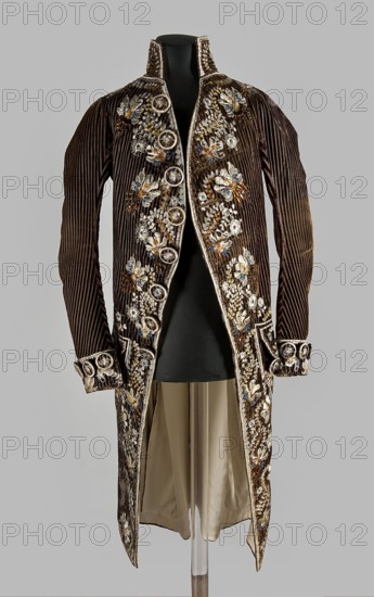 Habit à la française, tailcoat, floral ornaments, clothing, men's clothing, men's fashion, 18th century clothing. Habit of striped brown velvet, embroidered with fanciful flowers with large stamens and ears of grass. Fitted at the top, widened by pleats at the bottom, at the back, from the waist down. Long, set-in sleeves, cut in an arc at elbow height, finished with cuffs. Fastened with buttons, the skirts are cut in an arc, overlapping at the chest. Embroidery along the edges of the pockets, on the cuffs, pocket flaps, collar, and back pleats. The back is cut with a center seam; the back pleats do not overlap. Three buttons under the pocket flaps and on the cuffs. 1780s, Silesia, garment, length 111 cm, A long, richly decorated coat displayed on a black mannequin, made of dark brown ribbed velvet with vertical ridges and a lighter beige or cream inner lining visible through the front opening and hem; the coat has elaborate floral embroidery and embellishment in colors including white...