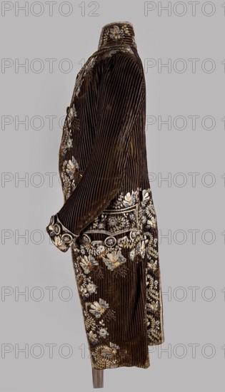 Habit à la française, tailcoat, floral ornaments, clothing, men's clothing, men's fashion, 18th century clothing. Habit of striped brown velvet, embroidered with fanciful flowers with large stamens and ears of grass. Fitted at the top, widened by pleats at the bottom, at the back, from the waist down. Long, set-in sleeves, cut in an arc at elbow height, finished with cuffs. Fastened with buttons; the skirts are cut in an arc, overlapping at the chest. Embroidery along the edges of the pockets, on the cuffs, pocket flaps, collar, and back pleats. The back is cut with a center seam; the back pleats do not overlap. Three buttons under the pocket flaps and on the cuffs. 1780s, Silesia, garment, length 111 cm., A side view of a long, dark brown velvet coat with vertical ribbed texture, decorated with embroidered floral motifs in colors including white, cream, blue, and gold; the coat has a high collar and long sleeves, ornate cuffs, and a waist area embellished with circular decorative elem...