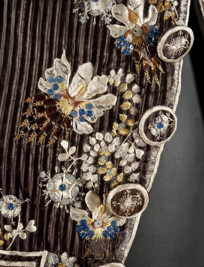 Habit à la française, tailcoat, floral ornaments, clothing, men's clothing, men's fashion, 18th century clothing. Habit of striped brown velvet, embroidered with fanciful flowers with large stamens and ears of grass. Fitted at the top, widened by pleats at the bottom, at the back, from the waist down. Long, set-in sleeves, cut in an arc at elbow height, finished with cuffs. Fastened with buttons, the skirts are cut in an arc, overlapping at the chest. Embroidery along the edges of the pockets, on the cuffs, pocket flaps, collar, and back pleats. The back is cut with a center seam; the back pleats do not overlap. Three buttons under the pocket flaps and on the cuffs. 1780s, Silesia, garment, length 111 cm, A close-up of a textured fabric panel with vertical ridges in deep brown, decorated with embroidered floral motifs in cream, white, gold, and pale yellow, accented by groups of small blue bead-like embellishments and faceted brown and amber beads, with circular embroidered appliqués e...