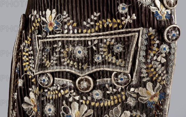 Habit à la française, tailcoat, floral ornaments, clothing, men's clothing, men's fashion, 18th century clothing. Habit of striped brown velvet, embroidered with fanciful flowers with large stamens and ears of grass. Fitted at the top, widened by pleats at the bottom, at the back, from the waist down. Long, set-in sleeves, cut in an arc at elbow height, finished with cuffs. Fastened with buttons, the skirts are cut in an arc, overlapping at the chest. Embroidery along the edges of the pockets, on the cuffs, pocket flaps, collar, and back pleats. The back is cut with a center seam; the back pleats do not overlap. Three buttons under the pocket flaps and on the cuffs. 1780s, Silesia, garment, length 111 cm, A section of dark brown ribbed fabric with ornate embroidery and embellishments, showing a decorative flap with rounded ornamental buttons and a pocket-like trim; the embroidery includes floral and leafy motifs with threadwork in ivory, pale beige, and yellow, and small accents in blu...
