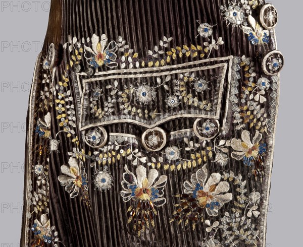 Habit à la française, tailcoat, repossession provenance noted, floral ornaments, men's clothing, men's fashion, 18th century clothing. Habit of striped brown velvet, embroidered with fanciful flowers with large stamens and ears of grass. Fitted at the top, widened by pleats at the bottom at the back from the waist down. Long set-in sleeves, cut in an arc at elbow height, finished with cuffs. Fastened with buttons; the skirts are cut in an arc, overlapping at the chest. Embroidery along the edges of the pockets, on the cuffs, pocket flaps, collar, and back pleats. The back is cut with a center seam; the back pleats do not overlap. Three buttons under the pocket flaps and on the cuffs. 1780s, Silesia, garment, length 111 cm., A close-up of a dark brown ribbed fabric panel with ornate embroidered floral motifs and decorative trim; there is a rectangular flap outlined with light-colored cord and circular button-like decorations, and the surrounding surface shows a group of embroidered flow...