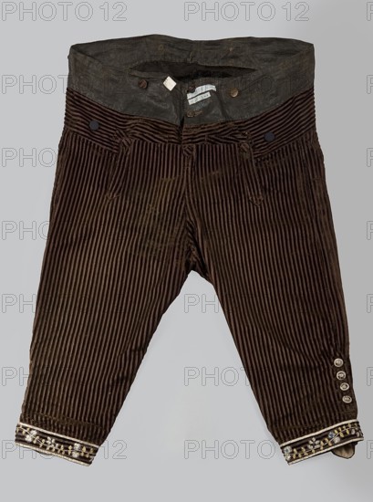 Trousers, short to the knee with a lapel, made of striped brown velvet; in the waist sewn into a wide belt of impregnated brown canvas. Lapel fastens with three covered buttons. Legs have slits fastened with five buttons each 1.5 cm diameter and finished with narrow embroidered cuffs. Additional metal and covered buttons for suspenders sewn on the belt. At the back, below the canvas belt, a triangular gusset of dark brown smooth velvet is sewn on. Men's clothing, floral ornaments, 1780s, Silesia; length 76 cm., A pair of dark brown corduroy pants with vertical ridges and a wide waistband in a darker grayish-brown fabric, featuring a few round buttons at the waist and a few belt loops; the pants have a tapered leg and a few decorative round buttons on one lower leg, and the cuffs are trimmed with embroidered bands showing floral-like motifs in light beige and gold tones; background is a plain light gray.