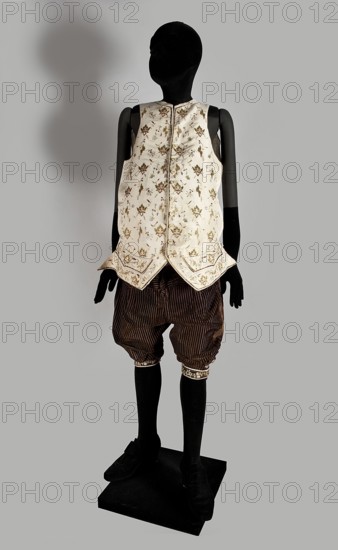 Trousers, trousers, clothing, floral ornaments, men's clothing, men's fashion, 18th century clothing, Trousers, short to the knee, with a lapel, made of striped brown velvet. In the waist, sewn into a wide belt of impregnated brown canvas. The lapel fastens with three covered buttons. The legs have slits, fastened with five buttons each one and a half centimeters in diameter, and finished with narrow, embroidered cuffs. Additional metal and covered buttons for suspenders are sewn on the belt. At the back, below the canvas belt, a triangular gusset made of dark brown smooth velvet is sewn on., 1780s, Silesia, garment, length 76 cm, A black mannequin stands on a black square base wearing a sleeveless cream-colored vest with a floral pattern in shades of gold and light brown, the vest having a pointed hem with decorative trim, paired with dark brown striped knee-length breeches with gathered cuffs that feature a narrow decorative band, and black stockings and black shoes; the background i...