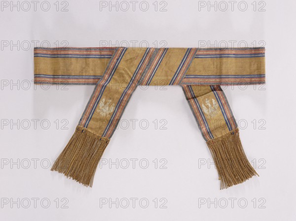 Textile belt, 19th century, Kraków. Belt made of solid fabric with the Polish Eagle at the ends. Sewn on morocco leather, fastened at the back, imitation lacing at the front. Length 86 cm, width 7.5 cm. Classified as a kontusz belt — a men's clothing/fashion accessory men's clothing accessories, national costume, Polish clothing., provenance: purchase., A woven sash laid flat against a pale background, featuring a golden base color with stripes in blue and red along its length, a few diagonal sections, and a group of long golden fringes at each end.