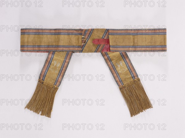 Textile belt, 19th century, Kraków. Belt made of solid fabric with the Polish Eagle at the ends. Sewn on morocco leather, fastened at the back, imitation lacing at the front. Length 86 cm, width 7.5 cm. Keywords: belts, Polish clothing, kontusz belt, fashion accessory, men's clothing accessory, eagles, national costume, Poland., A woven textile band laid flat on a pale background, composed of a horizontal section with two long vertical tails hanging down, each tail ending in a fringe; the fabric is primarily gold with woven narrow stripes in blue and red running along the edges and diagonally where pieces overlap, a small patch of muted red visible near the center where layers meet, and a few areas of lighter wear on the tails showing pale irregular marks.