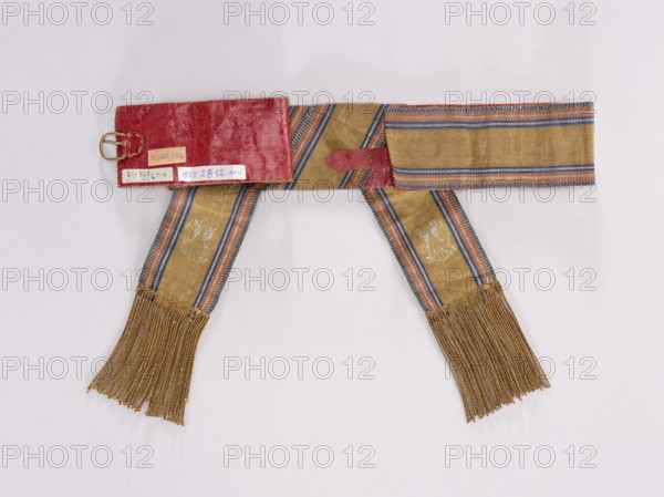 Textile belt, 19th century, Kraków. Belt made of solid fabric with the Polish Eagle at the ends; sewn on morocco leather, fastened at the back, imitation lacing at the front. Length 86 cm, width 7.5 cm. Associated with Polish clothing: kontusz belts, men's clothing accessories, national costume/fashion accessory; related to 1863–1864 examples., A textile sash laid flat on a pale background, consisting of a central band and two hanging ends with fringes; the sash fabric is primarily mustard yellow with woven stripes in blue and red along the edges and center, and there are areas of faded or worn lighter patches on the hanging sections; one end has a rectangular red leather or fabric piece attached with a metal buckle and a short red strap, and the red piece shows a slightly glossy, textured surface; the hanging ends finish with a group of long, gold-colored fringes.