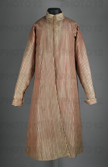 Zupan, Made of pink-carnassine silk with black stripes. Long, fitted at the top, flared at the bottom, with slightly overlapping skirts. Partially concealed fastening to the neck with small haberdashery buttons four visible at the collar. High, standing collar, adjustable at the back. Long, set-in sleeves with turn-up cuffs fastened with hooks. Side slit pockets. The back is cut like a kontusz, with a central column of fabric, widened from the waist on the sides by flared pleats, and laced at the top. The garment is lined with fustian up to the waist. 1780s, Poland. Garment length 120 cm, bottom circumference 282 cm., A long, striped coat displayed on a black mannequin form against a gray background, with a high stand-up collar and long sleeves; the coat features vertical narrow dark stripes on a pale pinkish-beige fabric, with the stripes continuing across the sleeves and cuffs and a visible front overlap.