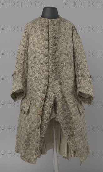 Szustokor, Szustokor made of silver lame with a net ornament featuring a pattern of small flowers. Fitted at the top, widened at the back from hip level down on the sides by folded pleats. Fastened at the front to the neck with seventeen buttons, without a collar. The lapels have curved edges. Long sleeves, set in at elbow height, cut in a curved manner, with wide, turned-up cuffs. The back is cut with a center seam, slit from the hip line, and the flaps do not overlap, decorated with silver braiding at the hem. Fabric-covered buttons are decoratively sewn on the cuffs and under the pocket flaps, four each, and two at the top of the side pleats. Men's garment, 1750–1775, Silesia. Length 108 cm., A long ornate coat displayed on a black mannequin neck, patterned all over with a floral brocade in muted green and beige tones with hints of pale pink, featuring a double-breasted front with a row of fabric-covered buttons, wide sleeves with buttoned cuffs, decorative buttoned flaps near the h...