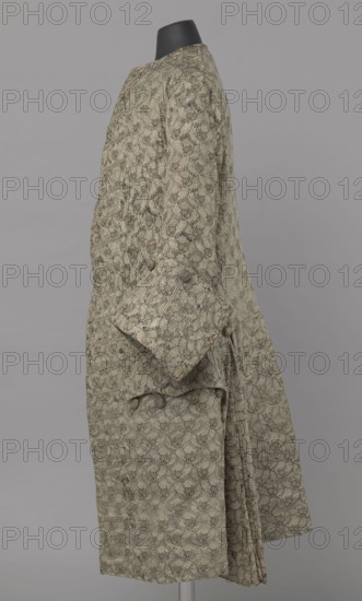 Szustokor, Szustokor made of silver lamé with a net ornament featuring a pattern of small flowers. Fitted at the top, widened at the back from hip level down on the sides by folded pleats. Fastened at the front to the neck with seventeen buttons, without a collar. The lapels have curved edges. Long sleeves, set in at elbow height, cut in a curved manner, with wide, turned-up cuffs. The back is cut with a center seam, slit from the hip line, and the flaps do not overlap, decorated with silver braiding at the hem. Fabric-covered buttons are decoratively sewn on the cuffs and under the pocket flaps, four each, and two at the top of the side pleats. 1750–1775. Silesia. Garment; length 108 cm., A side view of a long patterned coat displayed on a black mannequin head against a neutral gray background, the coat showing a repeated floral-like allover motif in muted green with accents of darker green and subtle brown, a few rounded fabric-covered buttons on the cuff and near the waist, a folded...