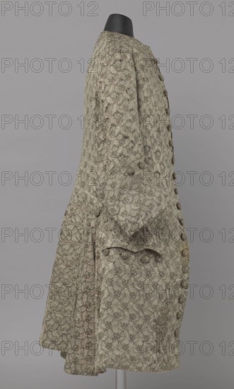 Szustokor, Szustokor made of silver lamé with a net ornament featuring a pattern of small flowers. Fitted at the top, widened at the back from hip level down on the sides by folded pleats. Fastened at the front to the neck with seventeen buttons, without a collar. Lapels have curved edges. Long sleeves set in at elbow height, cut in a curved manner, with wide, turned-up cuffs. Back cut with a center seam, slit from the hip line, flaps do not overlap, decorated with silver braiding at the hem. Fabric-covered buttons are decoratively sewn on the cuffs and under the pocket flaps, four each, and two at the top of the side pleats. Men's garment, 1750–1775, Silesia. Length 108 cm., A side view of a long patterned coat displayed on a black mannequin head against a neutral gray background, the coat showing an allover floral-like textured pattern in shades of pale green, beige, muted brown, and hints of darker accents, with a row of fabric-covered buttons along the front, a cuffed sleeve with d...
