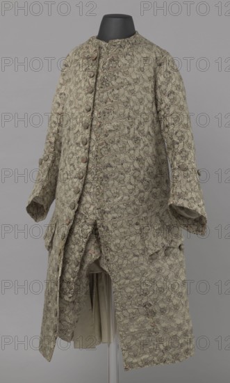 Szustokor, Szustokor made of silver lame with a net ornament featuring a pattern of small flowers. Fitted at the top, widened at the back from hip level down on the sides by folded pleats. Fastened at the front to the neck with seventeen buttons, without a collar. The lapels have curved edges. Long sleeves, set in at elbow height, cut in a curved manner, with wide, turned-up cuffs. The back is cut with a center seam, slit from the hip line, and the flaps do not overlap, decorated with silver braiding at the hem. Fabric-covered buttons are decoratively sewn on the cuffs and under the pocket flaps, four each, and two at the top of the side pleats. Men's garment, 1750–1775, Silesia. Length 108 cm., A long patterned coat displayed on a headless black mannequin, made of textured fabric in muted green with accents of darker green and small touches of pale pink and beige, featuring a high round neckline, a front opening with a vertical row of covered buttons on one side and decorative buttonh...