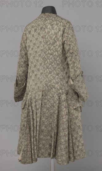 Szustokor, Szustokor made of silver lame with a net ornament featuring a pattern of small flowers. Fitted at the top, widened at the back from hip level down on the sides by folded pleats. Fastened at the front to the neck with seventeen buttons, without a collar. The lapels have curved edges. Long sleeves, set in at elbow height, cut in a curved manner, with wide, turned-up cuffs. The back is cut with a center seam, slit from the hip line, and the flaps do not overlap, decorated with silver braiding at the hem. Fabric-covered buttons are decoratively sewn on the cuffs and under the pocket flaps, four each, and two at the top of the side pleats. Men's garment, 1750–1775, Silesia. Length 108 cm., A back view of a patterned long coat displayed on a black mannequin head against a neutral gray background, the coat showing a repeating delicate floral-like motif in muted green tones with accents of darker green and small touches of faded reddish-pink, the fabric appearing textured and woven;...