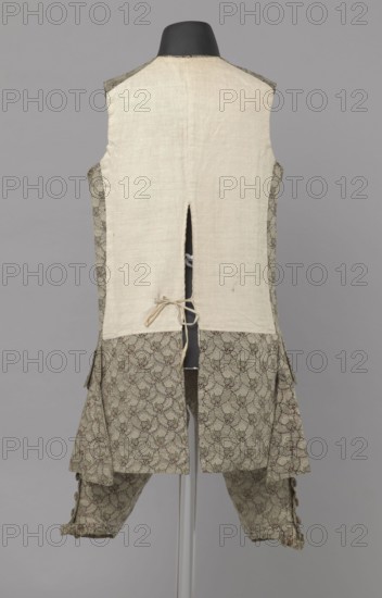 Szustokor, repossession provenance. Szustokor made of silver lamé with a net ornament featuring a pattern of small flowers. Fitted at the top, widened at the back from hip level down on the sides by folded pleats. Fastened at the front to the neck with seventeen buttons, without a collar. The lapels have curved edges. Long sleeves, set in at elbow height, cut in a curved manner, with wide, turned-up cuffs. The back is cut with a center seam, slit from the hip line, and the flaps do not overlap, decorated with silver braiding at the hem. Fabric-covered buttons are decoratively sewn on the cuffs and under the pocket flaps, four each, and two at the top of the side pleats. Men's garment, 1750–1775, Silesia. Length 108 cm., A back view of a sleeveless garment displayed on a black mannequin neck, with a plain light beige upper back panel and a patterned lower section and sides; the patterned fabric shows a repeating floral or vine-like motif in muted green and brown tones with small accents...