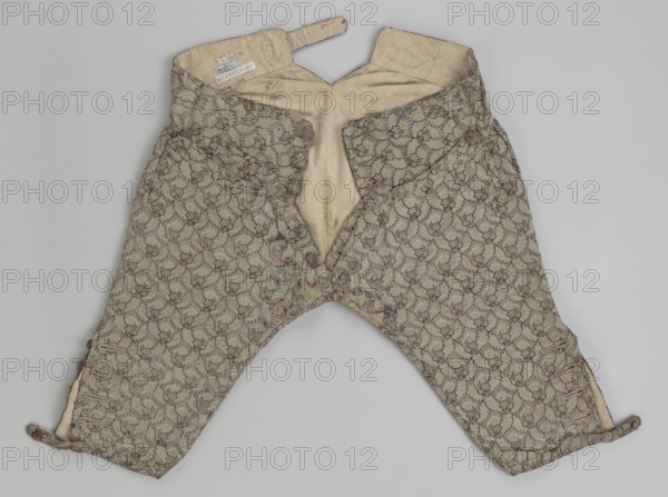 Short, knee-length men's trousers length 76 cm, 1750–1775, Silesia. Sewn into a wide belt with a buttoned fly, made of silver lamé with a net ornament forming a pattern of small flowers. Slit pockets on the sides of the fastener, closed with a side button. Belt is slit at the back and adjustable with a flap. Legs fasten at the bottom with four buttons and are finished with narrow cuffs. Floral ornaments., A pair of short trousers laid flat viewed from above, showing the inside and patterned outer fabric. The fabric has a repeating floral or leaf-like motif in muted tones, primarily beige with accents of green and small touches of muted red and dark brown. The interior lining is a plain light beige or cream color. There are a few visible buttons near the waistband and a group of buttonholes at the lower leg openings. The trousers show seams, stitching, and some areas of wear or fraying along the crotch and inner legs. The background is a uniform light gray.