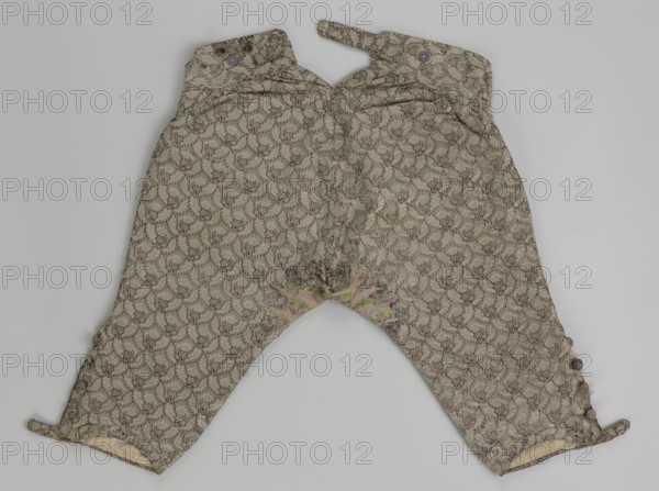 Short, knee-length men's trousers 1750–1775, Silesia. Sewn into a wide belt with a buttoned fly, made of silver lamé with a net ornament featuring a pattern of small flowers. Slit pockets at the sides of the fastener, fastened with a side button. Belt is slit at the back and adjustable with a flap. Legs fastened with four buttons at the bottom and finished with narrow cuffs. Length 76 cm., A pair of patterned pants laid flat against a light gray background, made from a beige fabric with a repeating floral-like motif in darker brown and muted green with small accents of faded pink, featuring a high waist with two straps extending from the top, a curved crotch seam with slight discoloration and wear, tapered legs with small rounded fabric-covered buttons along the lower outer seams, and visible inner lining in a pale off-white at the leg openings.
