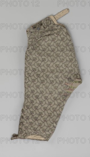 Trousers; short, knee-length trousers sewn into a wide belt, with a buttoned fly, made of silver lamé with a net ornament featuring a pattern of small flowers. Slit pockets on the sides of the fastener, fastened with a button on the side. The belt is slit at the back and adjustable with a flap. The legs are fastened with four buttons at the bottom and finished with narrow cuffs. Men's clothing, men's fashion; floral ornaments., Repossession, 1750–1775, Silesia. Length: 76 cm., A single fabric sleeve or pant leg laid flat, with a repeating floral-like woven pattern in muted tones of beige, gray, green, and faded pink, showing areas of wear and slight fraying along edges and seams; a small tab of plain beige fabric with stitching projects from the top, and a round darker button is visible near the upper edge; the lower edge shows a turned hem with an inner cream-colored lining peeking through, and a portion on one side has more pronounced wear revealing underlying layers with faded green...