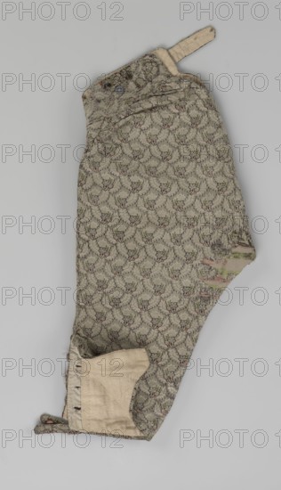 Short, knee-length men's trousers, sewn into a wide belt with a buttoned fly, made of silver lamé with a net ornament featuring a pattern of small flowers. Slit pockets on the sides of the fastener, fastened with a button on the side. The belt is slit at the back and adjustable with a flap. The legs are fastened with four buttons at the bottom and finished with narrow cuffs. Floral ornaments. Length 76 cm. Made c. 1750–1775, Silesia., A single fabric garment piece with an overall repeating floral-like pattern in muted green and beige, accented with small touches of pink and darker green, showing a curved shape and a strap at one end; a few areas reveal a plain beige lining and a small round button near the top, and there is visible wear with some fraying and a slightly discolored patch toward one edge.