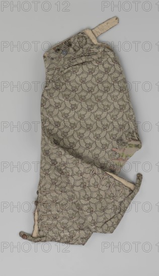 Trousers, short knee-length trousers sewn into a wide belt with a buttoned fly; made of silver lamé with a net ornament featuring a pattern of small flowers. Slit pockets at the sides of the fastener, fastened with a side button. Belt slit at the back and adjustable with a flap. Legs fastened with four buttons at the bottom and finished with narrow cuffs. Men's clothing, men's fashion. 1750–1775, Silesia. Length: 76 cm., A single patterned garment piece lies flat against a pale gray background, showing a textured fabric in muted beige-green with a repeating floral or scalloped motif in darker brown outlines and small touches of pink and olive green; the piece has curved and folded edges, visible buttonholes along one edge and a few small round buttons in a brownish tone, an attached fabric tab at one end in a lighter beige, and areas where the fabric appears worn or frayed with some irregular discoloration in soft pink and pale green.