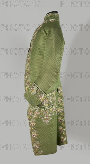 Habit à la française, Floral and plant embroidered motifs; green silk embroidered with small bouquets of flowers. Fitted at the top, widened at the back from the waist down with two slit pleats. Fastens at the front with hooks and eyes. Semicircularly cut lapels overlap at breast level and are cut off at the bottom. High standing collar. Long sleeves set in at elbow height, cut in an arc, finished with wide, slightly protruding cuffs. Side pockets at hip level covered with trapezoidal flaps. Back cut with a center seam; slits at hip level; center back flaps meet without a pleat. Embroidery runs in a wide band around the edges, cuffs, pockets, collar, and back pleats. Decorative buttons sewn in threes on the cuffs, under pocket flaps, two at the top of the back pleats, and in the pressed pleats. 1770s; Silesia place of origin / France place of creation. Garment; length 112 cm., A side view of a long green coat displayed on a form against a pale gray background, the coat made of a ribbed...