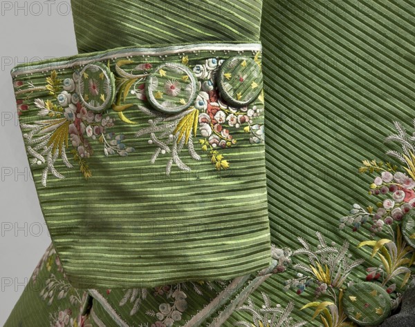 Habit à la française, Repossession provenance. Floral and plant motifs; fashion clothing: men's 18th-century garment. Green silk habit embroidered with small bouquets of flowers. Fitted at the top, widened at the back from the waist down with two slit pleats. Fastens at the front with hooks and eyes. Semicircularly cut lapels overlap at breast level and are cut off at the bottom. High standing collar. Long sleeves set in at elbow height, cut in an arc, finished with wide, slightly protruding cuffs. Side pockets at hip level covered with trapezoidal flaps. Back cut with a center seam; slits at hip level; center back flaps meet without a pleat. Embroidery in a wide band around edges, cuffs, pockets, collar, and back pleats. Decorative buttons sewn in threes on cuffs, under pocket flaps, two at top of back flaps, and in pressed pleats. 1770s, Silesia; France origin/creation. Garment length 112 cm., A close-up of a green textured fabric with diagonal ribbing and a few round fabric-covered ...