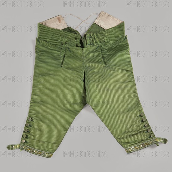 Trousers, short knee-length trousers with a flap, made of green silk fabric. Two small pockets under the flap, large pockets on the sides, and two small pockets for key rings above the flap. Legs end with narrow cuffs fastened at the sides with six embroidered buttons. Adjustable back, tied with a ribbon. Plant and floral motifs. 18th century 1770s. Silesia; made in France. Men's clothing; length 81 cm., A pair of short trousers made of green striped fabric laid flat against a gray background, showing a high waistband with inner white lining and tied strings, front waist tabs with fabric-covered buttons, front pockets, tapered legs that end above the ankle with a decorative trim, and buttoned cuffs with a few small round buttons on each side; visible colors include shades of green, white, gray, and a touch of metallic or shiny accents in the trim.