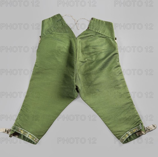 Trousers, plant motifs, floral motifs, clothing, fashion, men's clothing, 18th century clothing. Short, knee-length trousers with a flap, made of green silk fabric. Two small pockets under the flap. Large pockets on the sides. Two small pockets for key rings above the flap. The legs end with narrow cuffs, fastened at the sides with six embroidered buttons. Adjustable back, tied with a ribbon. 1770s, Silesia; made in France, length 81 cm., A pair of green pants laid flat on a light gray background, with a ribbed or pleated texture running horizontally across the fabric; the waistband area is slightly darker green with a folded-over appearance and has a tied cord at the top, while the legs taper toward the ankles and end with folded cuffs that reveal a patterned inner fabric in muted colors; the green fabric shows subtle variations in shade and some creasing along the seams.