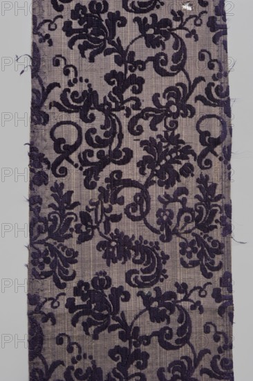 Fabric fragment, textiles, fragments of silk fabrics, patterned fabrics with plant motifs, cochineal and indigo dyes; fragments of a larger textile object. Patterned, looped purple velvet with plant motifs. Report motifs: plant tendril made of small, fanciful floral twigs in asymmetrical arrangements creating a net pattern. Weave construction: grosgrain background with silk bold and linen weft in a natural color, purple silk warp; looped silk purple cover. Dense plant composition, finely curved twigs with a variety of leaves and flowers – very delicate and with a refined, segmented pattern. Arranged in rows, with a half-report shift and with the calyx of the dominant flower tilted alternately to the left or right in the rows. Design report length 24.7 x width 13.2 cm. Attributed to Justyna Boninska, 17th century, Italy. Fabrics, height 110 cm, width 18 cm., A rectangular fabric panel with a woven ground in light gray and a raised ornate floral and scroll pattern in deep purple, showing...