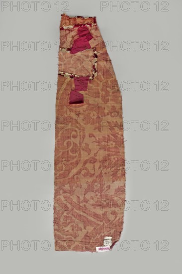 Fabric fragment, silk fabrics, fragments of patterned silk, chiseled velvet, Turkish fabrics. Beige and gold pattern fragment surrounded by cherry-cut fleece on a velvet background. Pattern features saz leaves with a fine floral ornament, lotus-shaped palmettes, and large winding forked thorny flower stems. 17th century, Ottoman Empire 1299–1922, fabrics. Height 86 cm, width 25.9 cm., A long narrow piece of patterned fabric with frayed edges laid flat on a light gray background, showing a faded floral and scroll design in shades of muted red and dusty pink with areas of lighter beige where the weave is worn, and a few irregular patches of deeper magenta fabric attached near the top; the textile surface has visible texture and small worn threads.